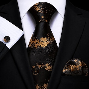 May include: A black necktie and matching pocket square featuring a gold floral pattern, worn with a white dress shirt, a black blazer, and a circular cufflink with a matching black and gold floral design on the white shirt cuff.