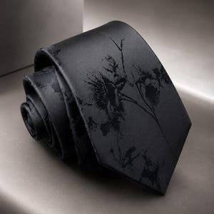 May include: A rolled black necktie featuring a subtle, dark-toned botanical print of branches and leaves. The fabric has a smooth, satin-like finish, and the tie is coiled to display the pointed end and the intricate, dark floral pattern across the surface.