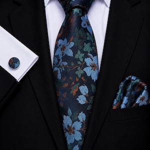 Könnte beinhalten: Eine dunkelblaue Krawatte mit einem Blumenmuster in Hellblau, Grün und Kupferbraun. Das Set enthält ein passendes Einstecktuch in der Tasche eines schwarzen Sakkos sowie einen runden Manschettenknopf an der Manschette eines weißen Hemdes.