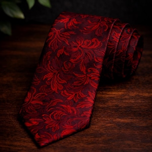 May include: A rolled burgundy tie with a repeating floral pattern in a darker red. The tie is made of textured fabric and is displayed on a dark wooden surface. The tie is a formal accessory.