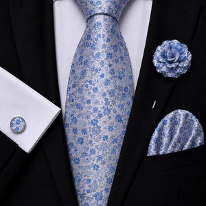 Op de afbeelding: Een zwart colbert gedragen met een wit overhemd, een lichtblauwe das met een klein bloemenpatroon, een bijpassend pochet, een blauwe bloemvormige reversspeld en een ronde manchetknop met een bijpassend bloemmotief op de witte manchet.