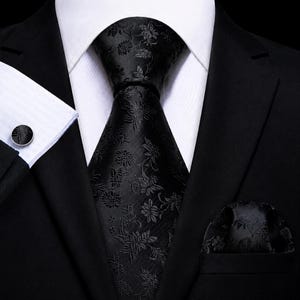 Könnte beinhalten: Eine schwarze Krawatte mit einem tonalen Blumenmuster, kombiniert mit einem passenden Einstecktuch und einem runden schwarzen Manschettenknopf. Das Set wird mit einem weißen Hemd und einem schwarzen Sakko zu einem formellen Outfit getragen.