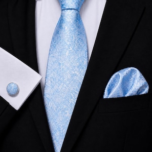 Könnte beinhalten: Eine hellblaue Krawatte mit dezentem Blumenmuster, ein passendes Einstecktuch und ein runder Manschettenknopf mit hellblauem Blumeneinsatz. Diese Artikel werden zu einem weißen Hemd und einem einfarbigen schwarzen Sakko getragen.