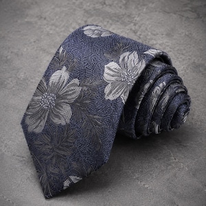 Puede incluir: Una corbata azul marino con un patrón floral tejido en tonos gris y plata. La tela tiene una apariencia texturizada y la corbata está enrollada en espiral, mostrando el extremo puntiagudo y el intrincado diseño botánico en toda la pieza.