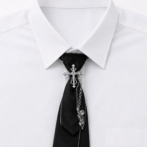 Może przedstawiać: Biała koszula z kołnierzykiem i gładki czarny krawat. Krawat ozdobiony jest srebrną metalową broszką w kształcie krzyża z osadzonymi przezroczystymi kryształkami oraz zwisającym łańcuszkiem, który opada po prawej stronie materiału.