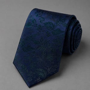 May include: A dark navy blue necktie featuring a tonal jacquard woven pattern of various leaves and floral elements. The fabric has a subtle sheen, and the tie is rolled at the top, displaying a pointed tip at the bottom end of the garment.
