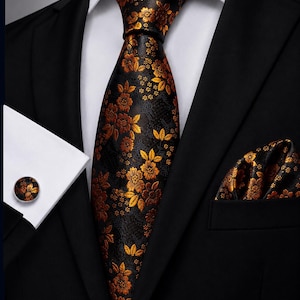 Può includere: Una cravatta nera e una pochette abbinata con un vivace motivo floreale arancione e dorato. Il set include un gemello circolare con lo stesso disegno floreale, il tutto indossato con una camicia bianca e una giacca da completo nera.