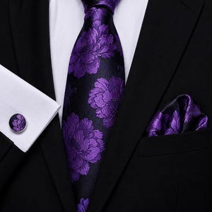Könnte beinhalten: Ein schwarzes Sakko getragen mit einem weißen Hemd, einer violetten Krawatte mit Blumenmuster, einem passenden Einstecktuch mit Blumenmuster und einem einzelnen runden Manschettenknopf mit demselben violetten Blumenmuster auf silberfarbenem Metall.