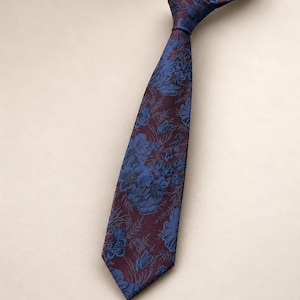 May include: A necktie featuring a deep burgundy background with a repeating pattern of blue floral motifs and foliage. The tie is tied in a knot at the top, displaying a pointed tip at the bottom and a textured fabric surface throughout the design.