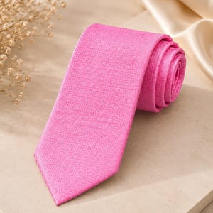 May include: A rolled-up, bright pink tie with a subtle geometric pattern. The tie is made of a smooth fabric and is displayed on a neutral-toned surface. The tie's vibrant colour and texture are highlighted in the image.