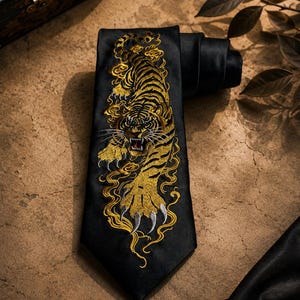 Könnte beinhalten: Schwarze Krawatte mit einem detaillierten, goldgestickten Tigerdesign. Der Tiger ist mit offenem Maul und ausgestreckten Krallen dargestellt, umgeben von goldenen, wolkenartigen Akzenten. Die Krawatte ist aus glattem, dunklem Stoff gefertigt.
