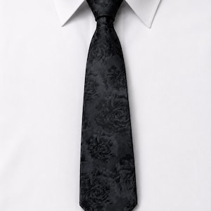 May include: A black necktie featuring a tonal floral pattern with a subtle sheen, tied in a standard knot and positioned against a crisp white collared dress shirt. The fabric displays a repeating rose motif in a matching black shade.