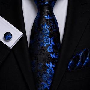 Può includere: Una cravatta nera con un vivace motivo floreale blu, abbinata a una pochette coordinata e a un gemello circolare. Il set è indossato con una camicia bianca e una giacca da abito nera, creando un insieme formale e ben coordinato.