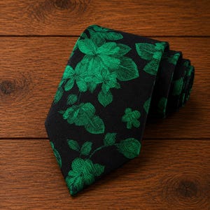 Peut inclure: Une cravate présentant un fond noir avec un motif floral vert vif. Le tissu affiche un design tissé détaillé de diverses fleurs et feuilles dispersées sur la surface, la cravate étant pliée dans une disposition en couches enroulées.