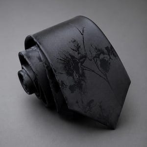 May include: A black necktie featuring a subtle, dark-toned floral pattern. The tie is rolled into a coil, showcasing the smooth fabric texture and the intricate, shadowy botanical print that extends across the surface of the garment.