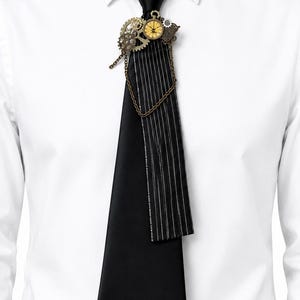 Può includere: Una cravatta nera con un pannello a righe verticali sul lato destro, decorata con un gruppo di ingranaggi color bronzo metallizzato, un piccolo quadrante circolare e una catena decorativa drappeggiata sul tessuto, indossata sopra una camicia bianca.
