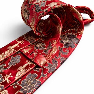 Pode incluir: Uma gravata vermelha com um padr&atilde;o floral e de p&aacute;ssaros ornamentado em ouro e preto. A gravata est&aacute; enrolada, mostrando o design detalhado. O tecido tem um leve brilho, real&ccedil;ando as cores ricas e os detalhes intrincados do padr&atilde;o.