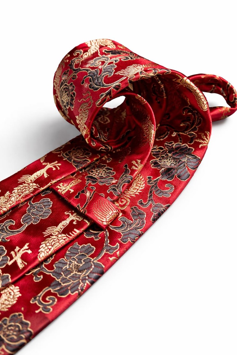 Pode incluir: Uma gravata vermelha com um padr&atilde;o floral e de p&aacute;ssaros ornamentado em ouro e preto. A gravata est&aacute; enrolada, mostrando o design detalhado. O tecido tem um leve brilho, real&ccedil;ando as cores ricas e os detalhes intrincados do padr&atilde;o.