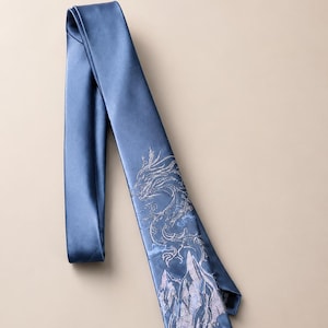 May include: A blue necktie featuring a detailed embroidered design of a dragon winding above mountain peaks. The fabric has a smooth, satin-like finish, and the embroidery is rendered in light, metallic-toned thread against the solid blue background.