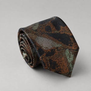 May include: A necktie rolled into a coil, featuring a textured pattern of abstract shapes in shades of black, copper, and muted sage green. The fabric has a woven appearance with a visible cross-hatch texture throughout the entire surface.