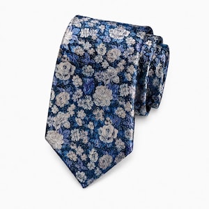 Könnte beinhalten: Eine Krawatte mit tiefblauem Hintergrund und einem dichten, gewebten Blumenmuster in Silber-, Hellblau- und Weißtönen. Der Stoff hat eine strukturierte Optik mit einer spitz zulaufenden Form und ist oben rechts umgeschlagen.