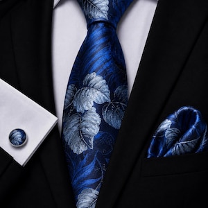 Könnte beinhalten: Ein Set aus einer blauen Krawatte, einem Einstecktuch und einem Manschettenknopf mit silbernem Blattmuster. Die Accessoires sind auf einem schwarzen Sakko und einem weißen Hemd arrangiert. Krawatte, Einstecktuch und Manschettenknopf zeigen dasselbe Design.