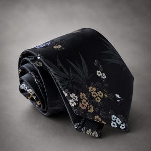 Puede incluir: Una corbata negra con un patrón tejido de flores blancas, doradas y azul claro junto con hojas de bambú. La corbata está enrollada en espiral, mostrando el intrincado diseño botánico sobre su superficie de tela oscura y lisa.