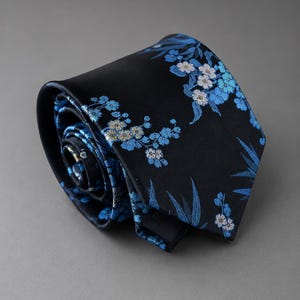 Könnte beinhalten: Eine schwarze Krawatte, die zu einer Rolle gewickelt ist und ein gewebtes Muster aus hellblauen und weißen Kirschblüten sowie schmalen Blättern aufweist. Der Stoff hat eine glatte, satinartige Oberfläche mit einem floralen Design.