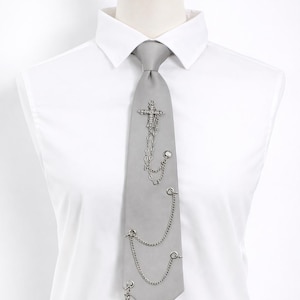 Peut inclure: Une cravate grise ornée de croix et de chaînes argentées. La cravate est présentée sur une chemise blanche et un mannequin. Les croix sont ornées de petits détails. Les chaînes pendent des croix et sont attachées à la cravate.