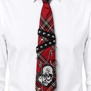 Peut inclure: Une cravate à motif tartan rouge ornée d'une bande noire cloutée sur la partie supérieure, d'un écusson brodé représentant un crâne blanc, d'épingles à nourrice argentées, de chaînes métalliques pendantes et de petits clous pyramidaux.