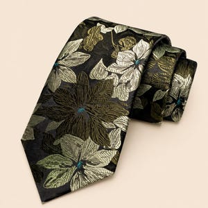 Könnte beinhalten: Eine Krawatte mit schwarzem Hintergrund und einem gewebten Blumenmuster in Olivgrün, Salbei und Metallic-Silber. Das Design zeigt große, detaillierte Blumenmotive mit kleinen, leuchtend türkisfarbenen Akzenten in der Mitte jeder Blüte.