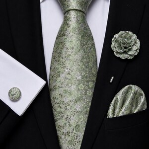 May include: A sage green necktie featuring a detailed floral pattern, paired with a matching pocket square, a round floral cufflink, and a floral lapel pin. These accessories are worn with a crisp white dress shirt and a black suit jacket.