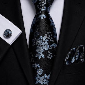 Könnte beinhalten: Eine schwarze Krawatte mit hellblauem Blumenmuster, kombiniert mit einem passenden Einstecktuch und einem runden Manschettenknopf. Das Set wird zu einem weißen Hemd und einem schwarzen Sakko getragen, was ein abgestimmtes formelles Outfit ergibt.