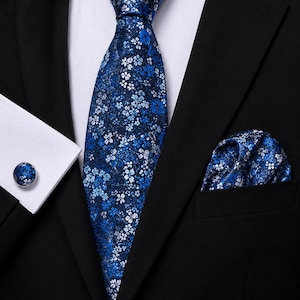 Op de afbeelding: Een blauwe das met een dicht patroon van kleine witte en lichtblauwe bloemen, gecombineerd met een bijpassend pochet en een enkele ronde manchetknop. De set wordt gedragen met een wit overhemd en een zwart colbert.