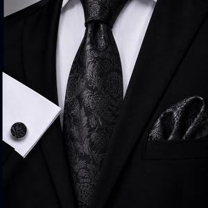 Könnte beinhalten: Eine schwarze Krawatte mit einem tonalen floralen Damastmuster, kombiniert mit einem passenden Einstecktuch und einem runden Manschettenknopf mit abgestimmtem Blumenmuster, getragen zu einem weißen Hemd und einem schwarzen Sakko.