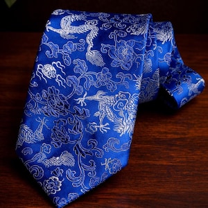 May include: A blue necktie with a detailed silver floral and dragon pattern. The tie is made of a shiny fabric and is folded over on a dark wooden surface. The design includes flowers, dragons, and other decorative elements.