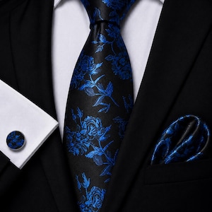 May include: A black necktie featuring a vibrant blue floral pattern, paired with a matching pocket square and a single round cufflink with the same blue floral design, all worn against a crisp white dress shirt and a black suit jacket.