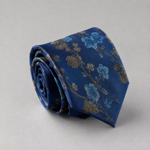 Peut inclure: Une cravate enroulée en tissu bleu marine profond ornée d'un motif répétitif de fleurs bleu clair et or métallique sur de fines tiges ramifiées. La cravate présente une finition satinée lisse et une extrémité pointue, présentée enroulée.