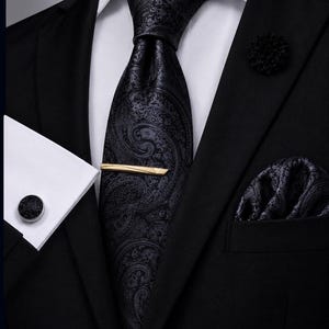 Könnte beinhalten: Ein schwarzes Sakko über einem weißen Hemd mit einer schwarzen Krawatte mit Paisleymuster, einer goldenen Krawattenklammer, einem passenden Einstecktuch, einer schwarzen Ansteckblume und einem schwarzen runden Manschettenknopf am Ärmel.