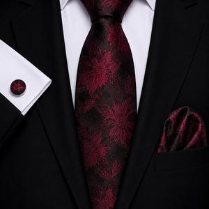 Può includere: Una giacca da abito nera indossata sopra una camicia bianca con una cravatta bordeaux scuro con motivo floreale. Il set include una pochette bordeaux abbinata con motivo floreale e un gemello circolare con lo stesso disegno floreale.