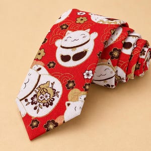 Könnte beinhalten: Eine rote Krawatte mit einem wiederkehrenden Muster aus weißen winkenden Katzen, bekannt als Maneki-neko, durchsetzt mit kleinen goldenen und schwarzen Blumenmotiven sowie roten wellenförmigen Linien. Der Stoff weist eine sichtbare Webstruktur auf.
