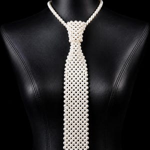 Puede incluir: Una corbata construida a partir de una cuadrícula de pequeñas perlas artificiales blancas y redondas. La corbata presenta un nudo estructurado en la parte superior y un cuerpo largo y rectangular que termina en punta, todo unido por un hilo fino.