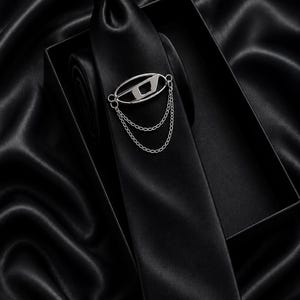 May include: A black satin tie with a silver-tone tie clip featuring a stylized eye design and dangling chains. The tie is displayed in a black box, set against a black, silky background.