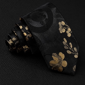 Könnte beinhalten: Eine schwarze Krawatte mit einem eingewebten Muster aus goldenen Blumenmotiven und kreisförmigen Designs. Der Stoff hat einen dezenten Glanz und die Krawatte ist zu einer geschichteten Rolle gewickelt, die den Kontrast zwischen dem dunklen Hintergrund und den goldenen Details betont.