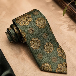 Può includere: Una cravatta verde arrotolata con un motivo floreale ripetuto in oro e marrone. La cravatta è realizzata in un tessuto strutturato e viene esposta su una superficie dai toni neutri. Il design floreale è dettagliato e intricato.