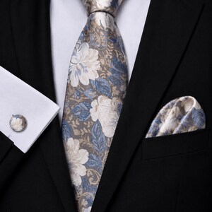 May include: A necktie, pocket square, and cufflink set featuring a floral pattern in shades of blue, cream, and taupe. The items are worn with a black suit jacket and a crisp white dress shirt, showcasing a coordinated design with large blossoms.