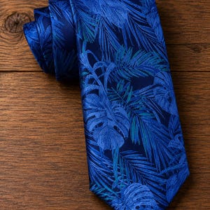 May include: A necktie featuring a deep blue base color with a repeating pattern of tropical palm and monstera leaves in shades of lighter blue and teal. The fabric has a subtle sheen, and the tie is folded at the top, showing a pointed end.