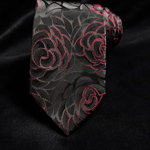 Könnte beinhalten: Eine Krawatte mit einem dunkelgrauen und schwarzen Blumenmuster, das mit feinen roten Stickereien umrandet ist. Der Stoff hat einen dezenten Glanz und das Design besteht aus sich wiederholenden rosenartigen Formen auf der gesamten Fläche.