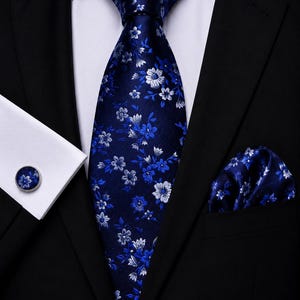 Peut inclure: Une cravate bleu foncé ornée d'un motif floral blanc et bleu clair, accompagnée d'une pochette assortie et d'un bouton de manchette rond présentant le même motif, le tout porté avec une chemise blanche et une veste de costume noire.
