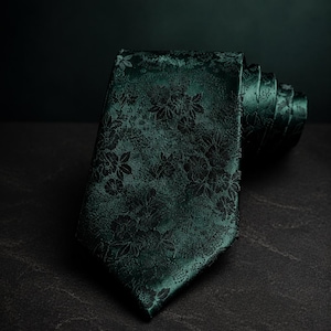 May include: A dark emerald green necktie featuring a woven black floral pattern. The fabric has a subtle sheen, and the tie is rolled at the end, displaying the intricate botanical design across the surface of the pointed blade and the rolled section.